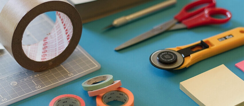 Close-up of crafting supplies including scissors, tape rolls, rotary cutter, and sticky notes on a blue surface.