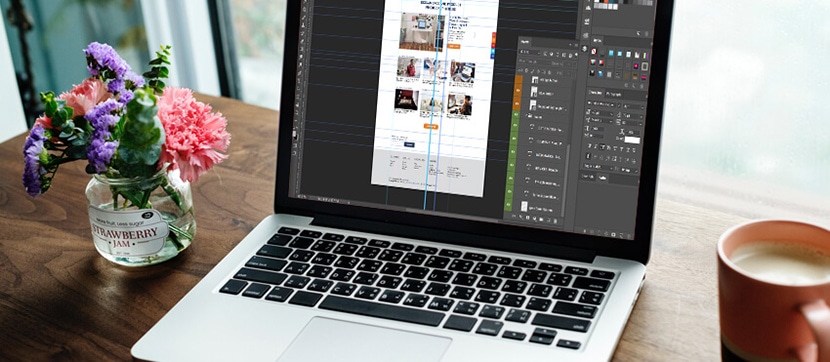 Open laptop displaying Adobe Photoshop design layout, beside flowers and a coffee cup on a wooden table.