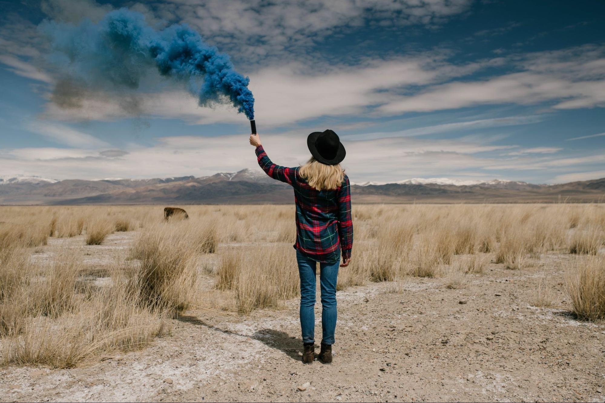 Person in hat and flannel holding blue smoke bomb in open desert field under cloudy sky