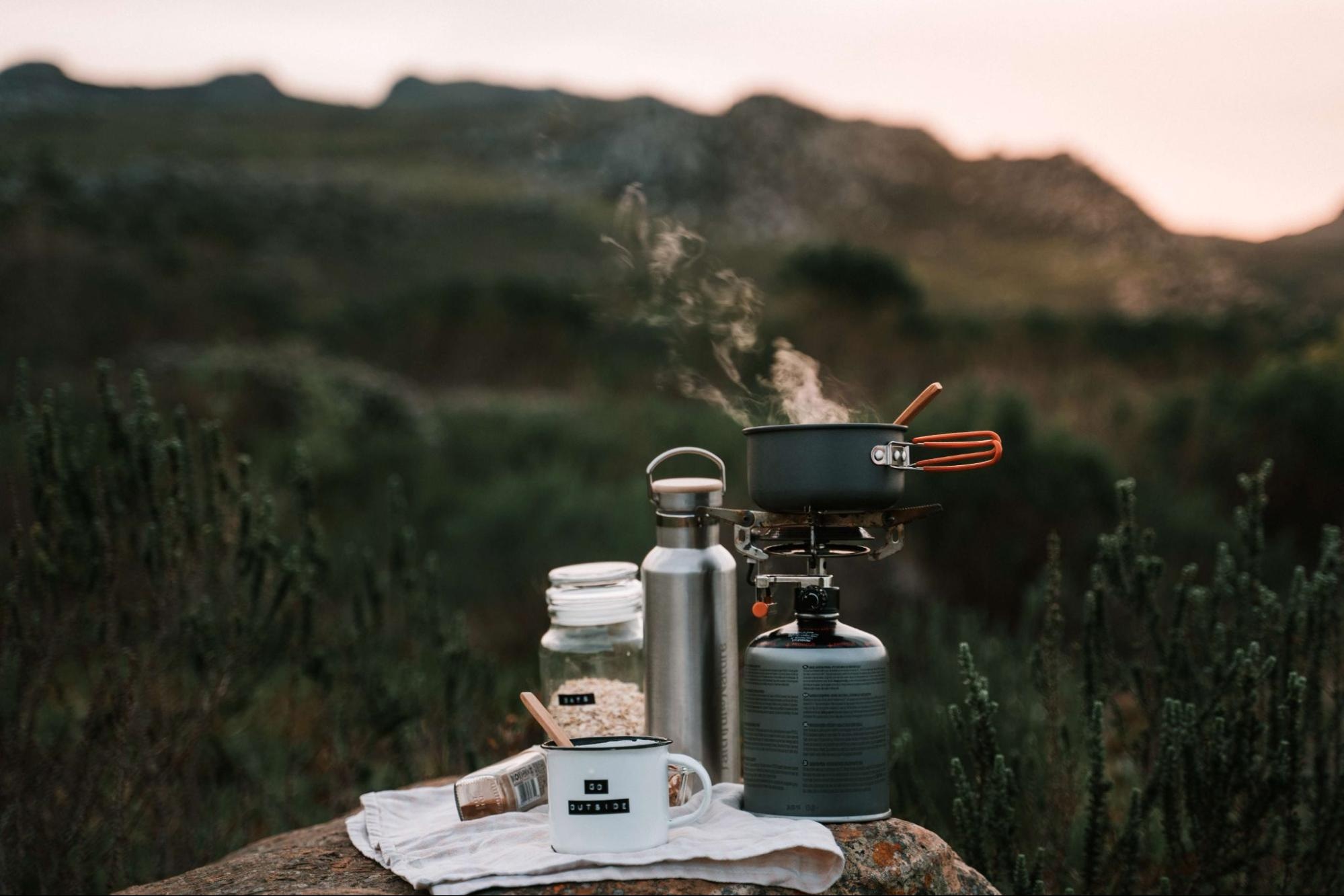 Camping cook setup with pot on stove, mug, and thermos in a scenic outdoor setting