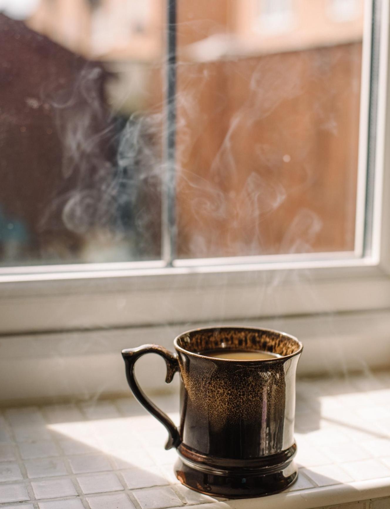 Brown ceramic mug with steam by a sunlit window, creating a warm and cozy atmosphere