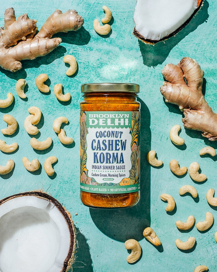 Brooklyn Delhi Coconut Cashew Korma simmer sauce jar surrounded by fresh ginger, cashew nuts, and coconut halves on a vibrant turquoise background.
