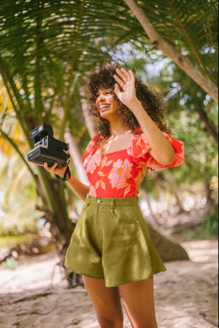 Smiling woman in floral top and olive shorts holding instant camera under palm trees