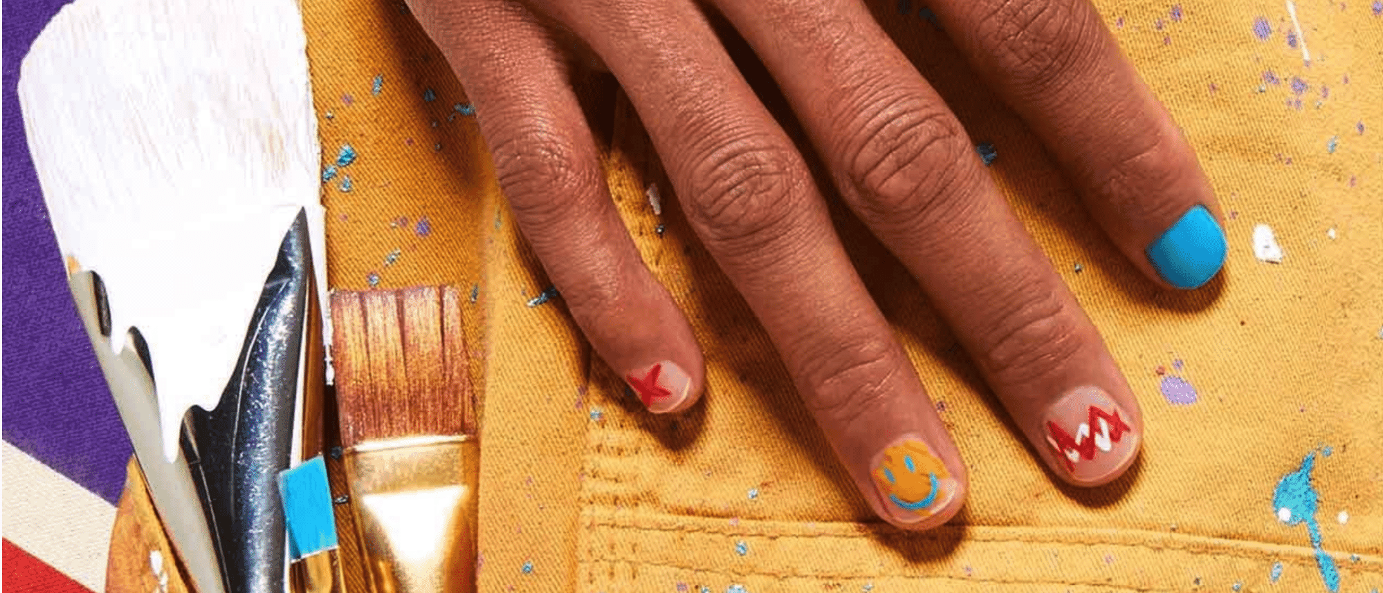 Close-up of hand with creative nail art, including smiley face and abstract designs, resting on paint-splattered yellow fabric with paintbrushes nearby.