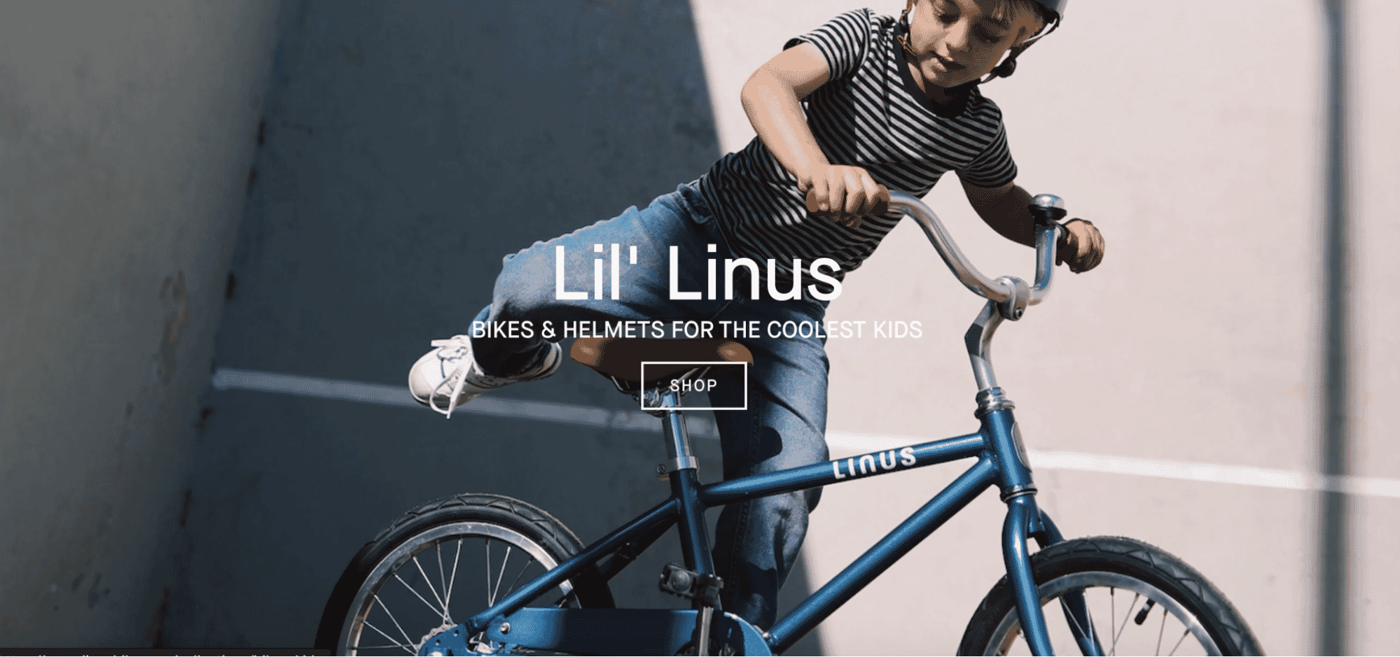 Child riding a blue Linus bike while wearing a helmet, captured mid-action against a concrete urban background with bold shadows.
