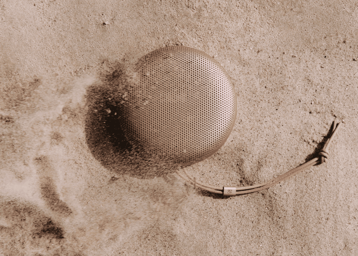 Rose gold Bang & Olufsen portable speaker partially buried in sand with leather strap, illustrating durability and beach-friendly audio design.