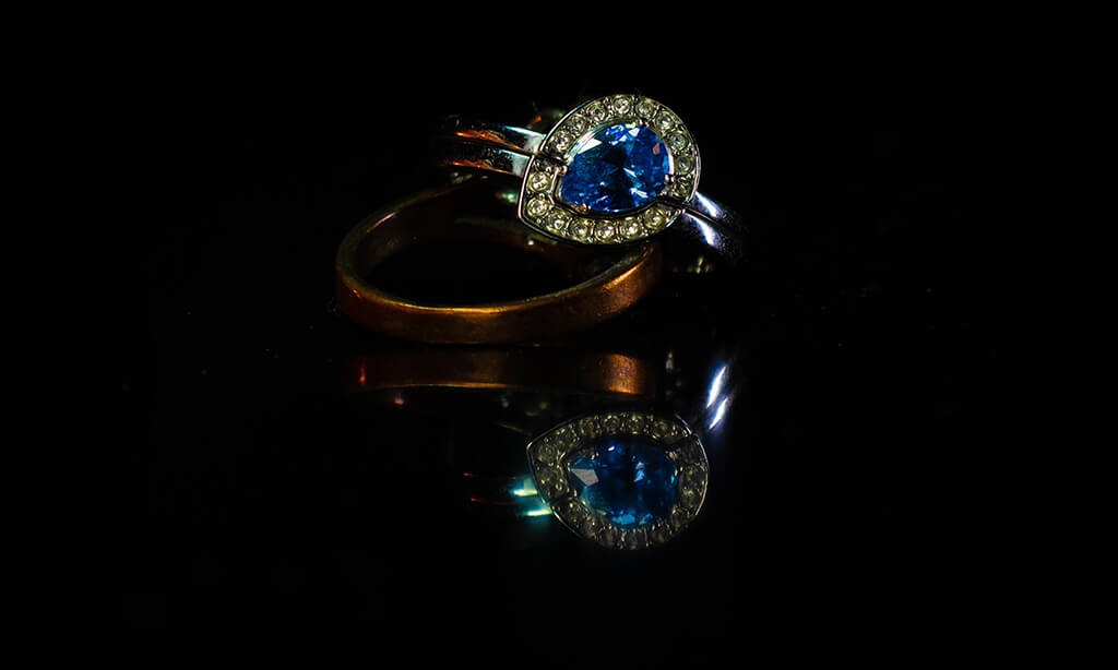 Elegant blue gemstone ring with diamond accents, reflected on a glossy black surface