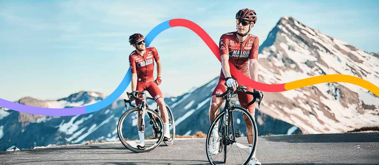 Two cyclists in red jerseys riding on a scenic mountain road with snow-covered peaks in the background. A colorful digital graphic overlay curves across the image, adding a dynamic visual effect.