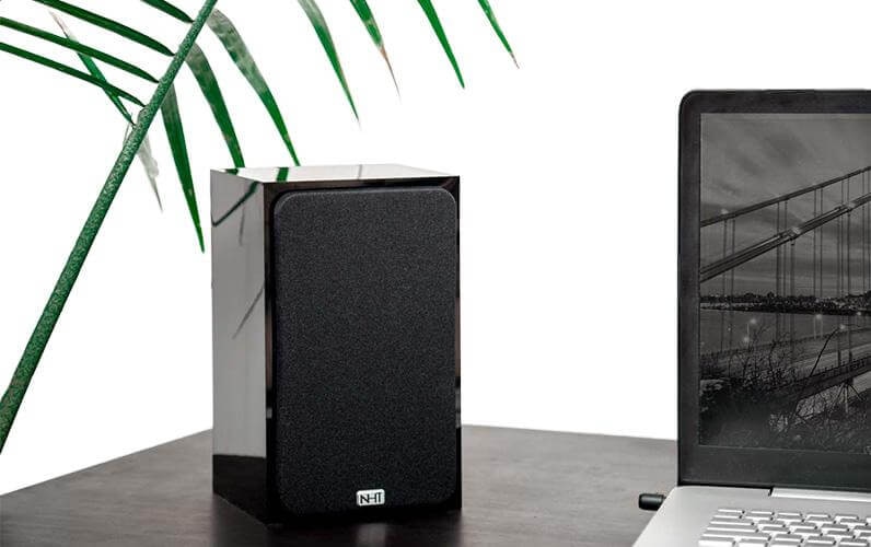 Minimalist speaker setup with a black NHT speaker, a laptop, and a clean, natural backdrop.