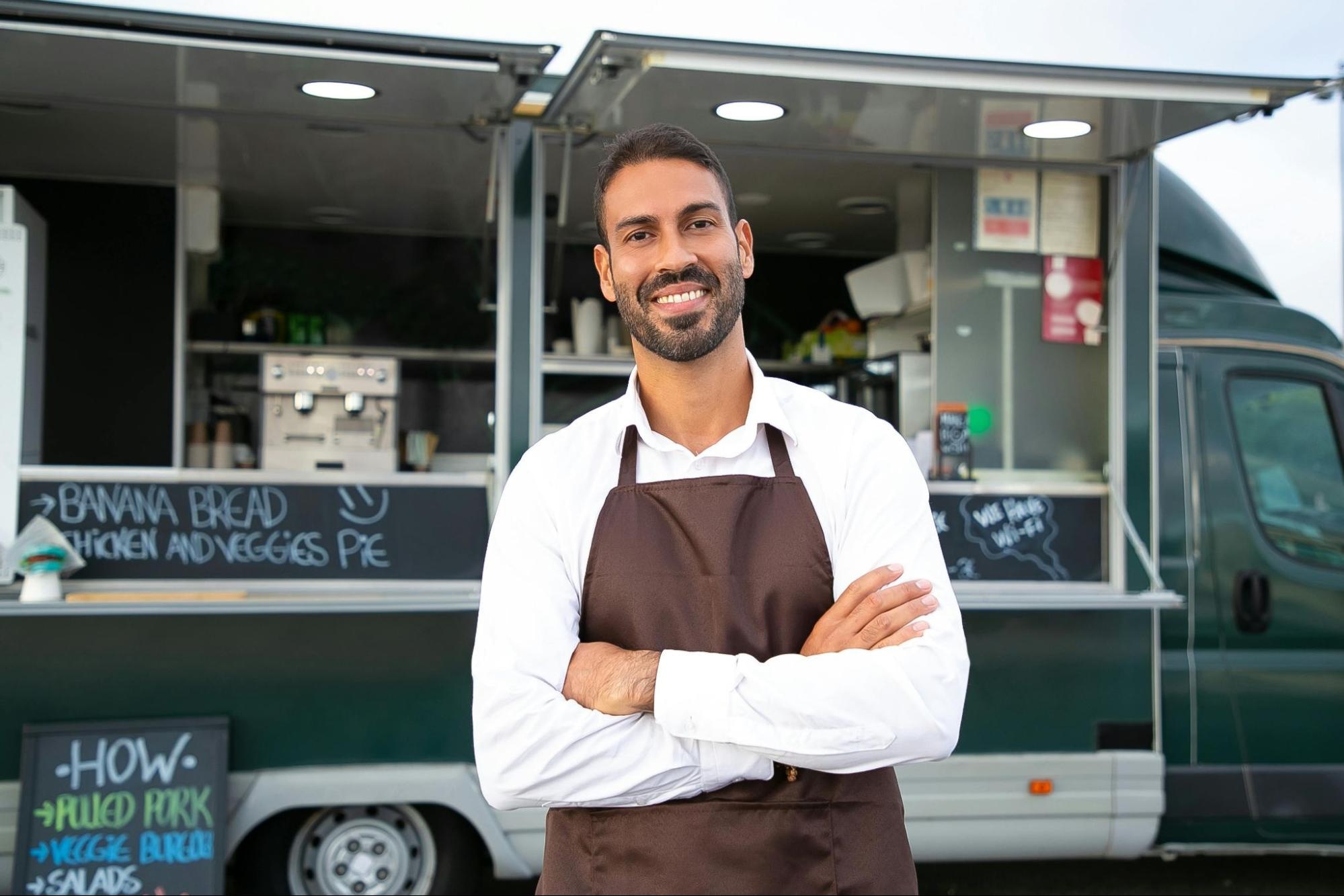 Smiling man in apron standing in front of his food truck with arms crossed