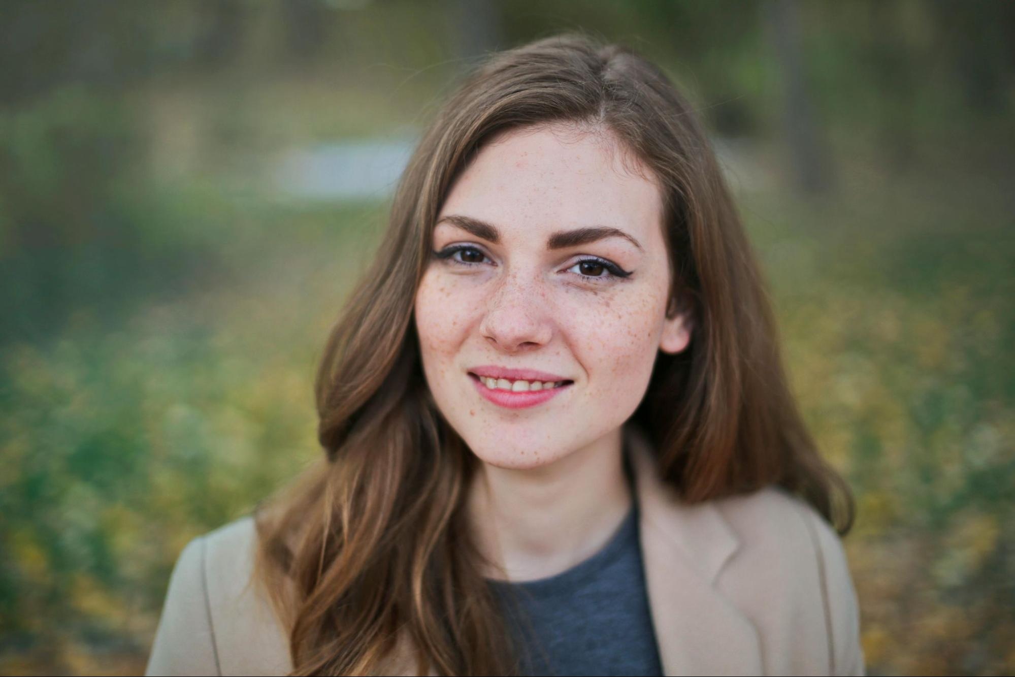 Young woman with light freckles and long hair smiling in an outdoor setting with soft, blurred background