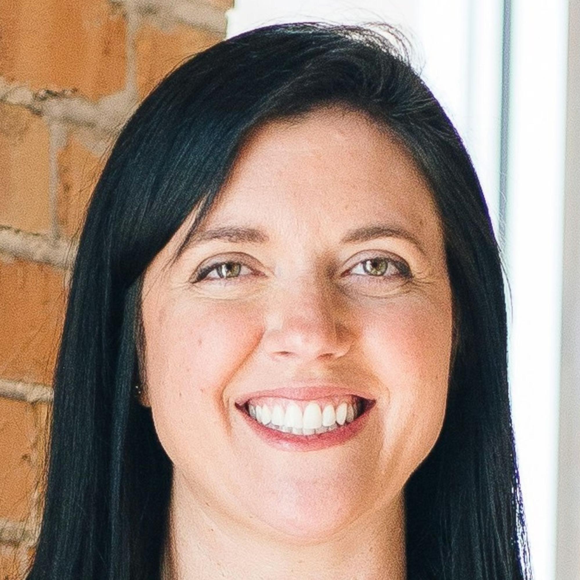 Square crop of a woman with straight black hair smiling in front of a sunlit brick wall inside an office.