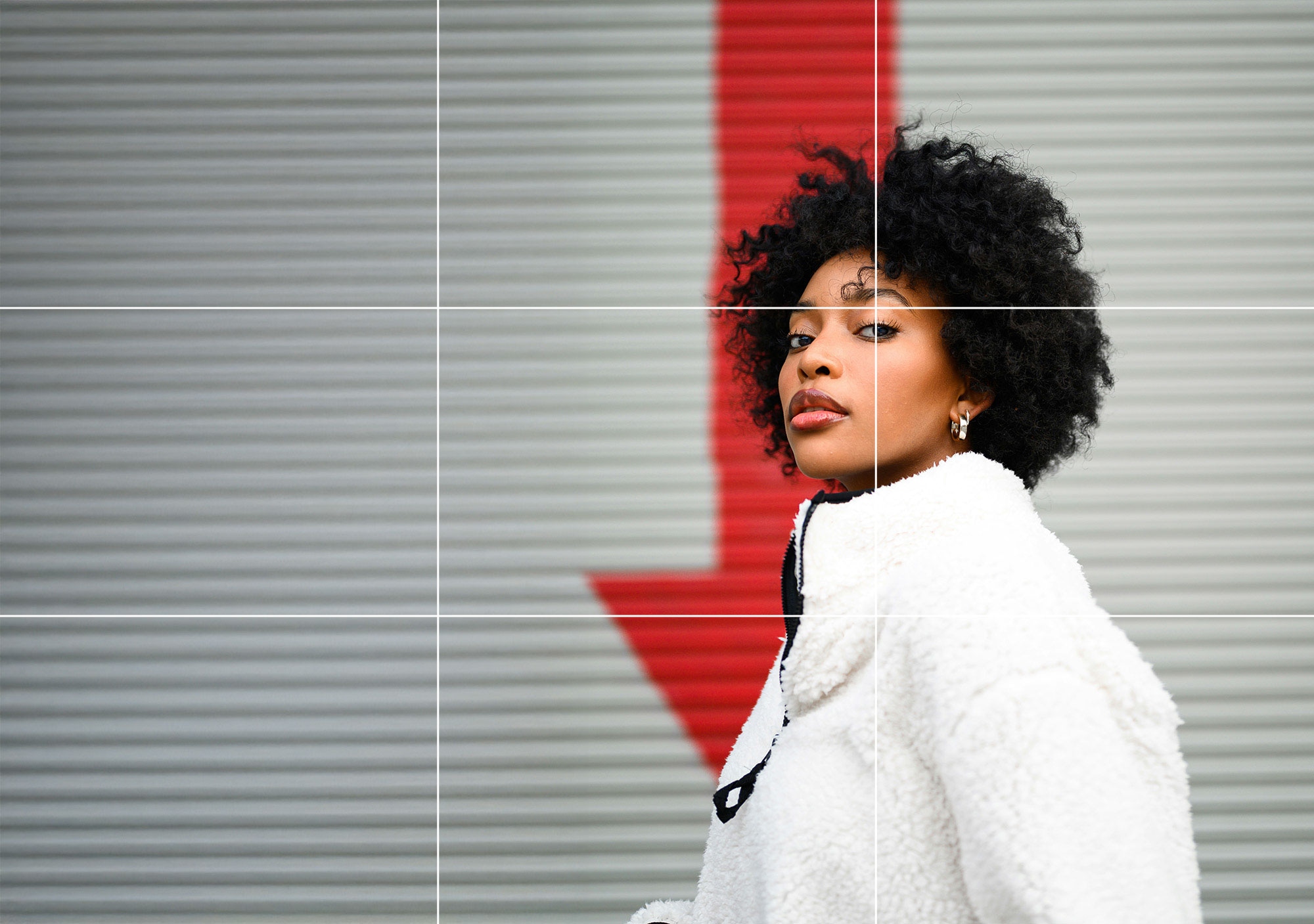 Confident woman in a white jacket posing in front of a corrugated wall with a bold red stripe in the background.