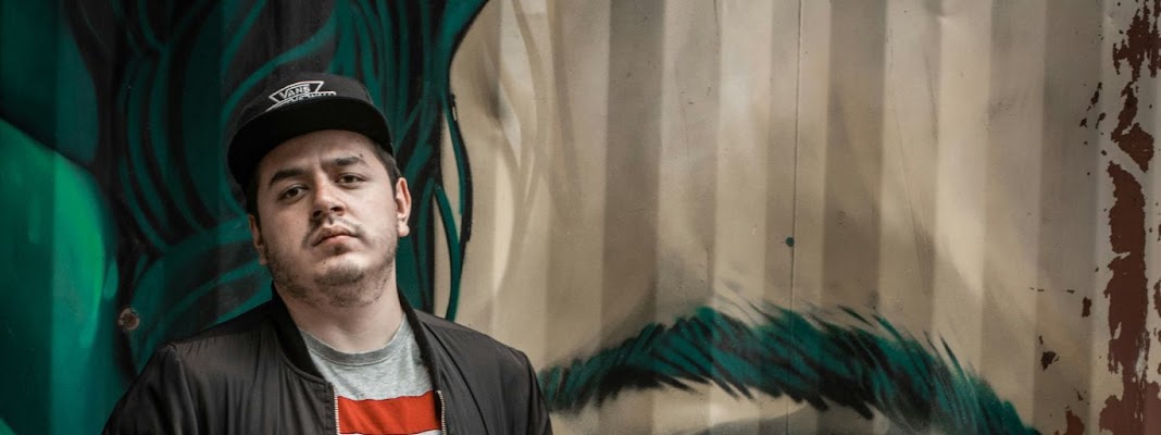 Panoramic crop of a man in a cap standing against a graffiti-covered wall with a large eye mural in the background.