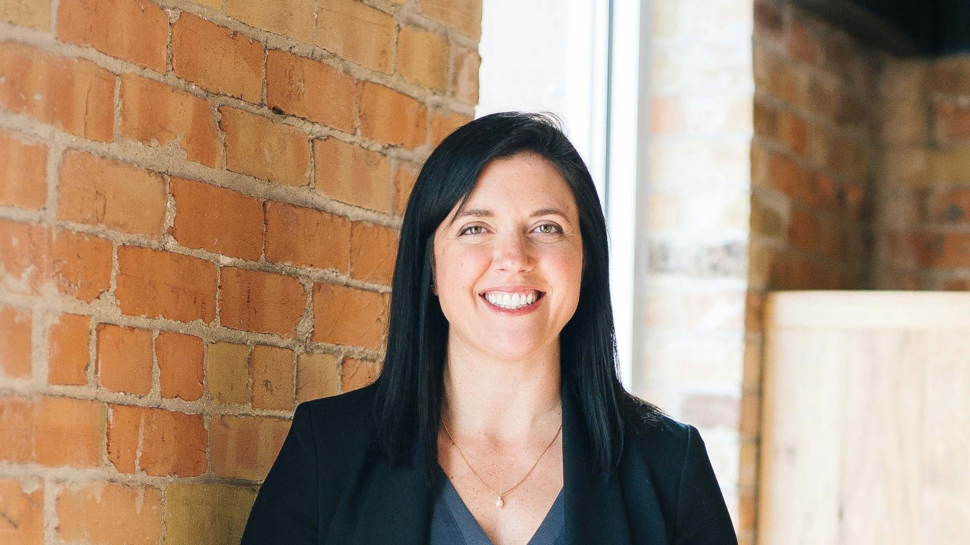 Widescreen crop of a woman in business attire smiling with arms relaxed, standing inside a brick-walled office with natural light.