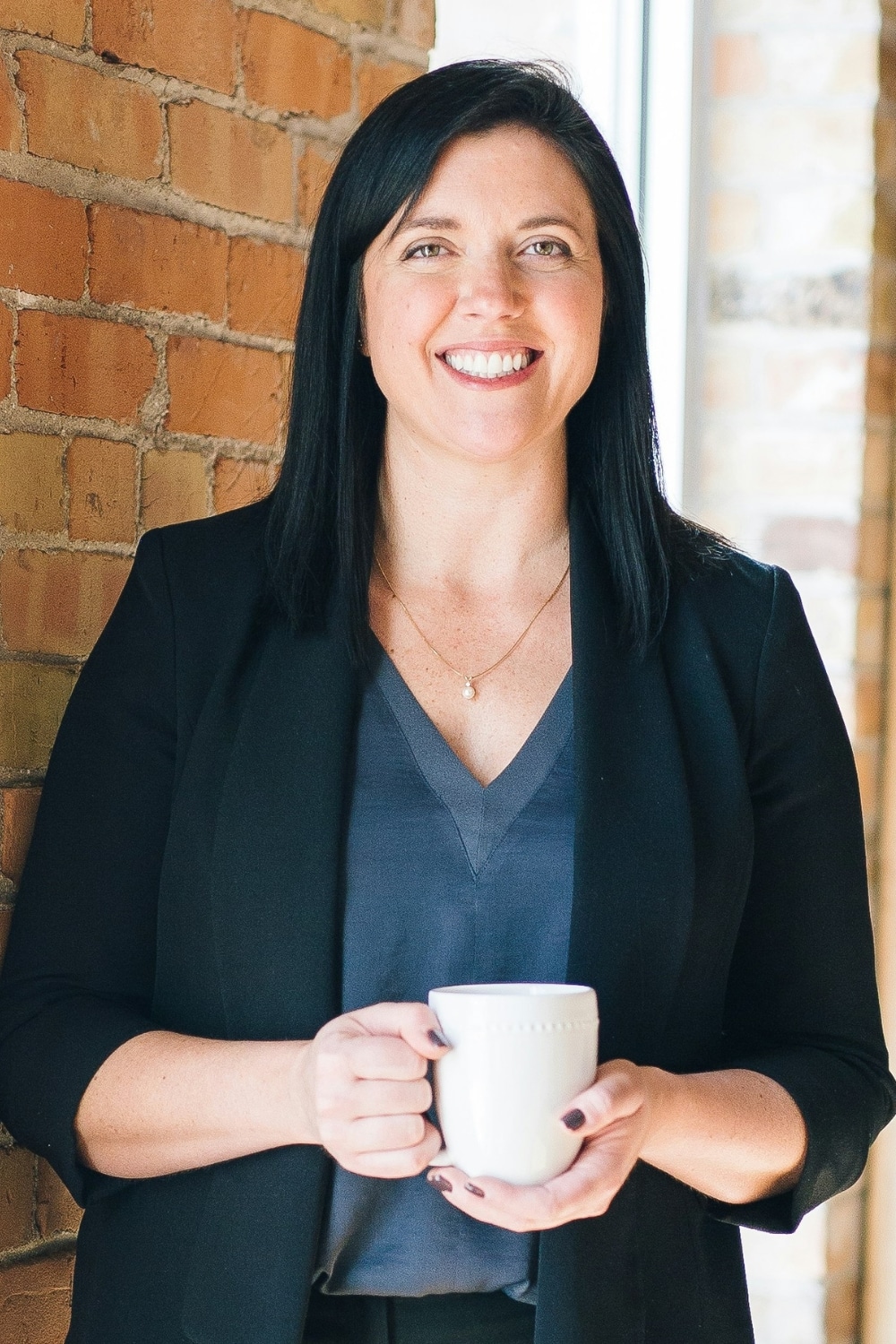 Classic photo print crop of a smiling woman holding a white mug, standing near a brick wall with soft daylight coming through the window.