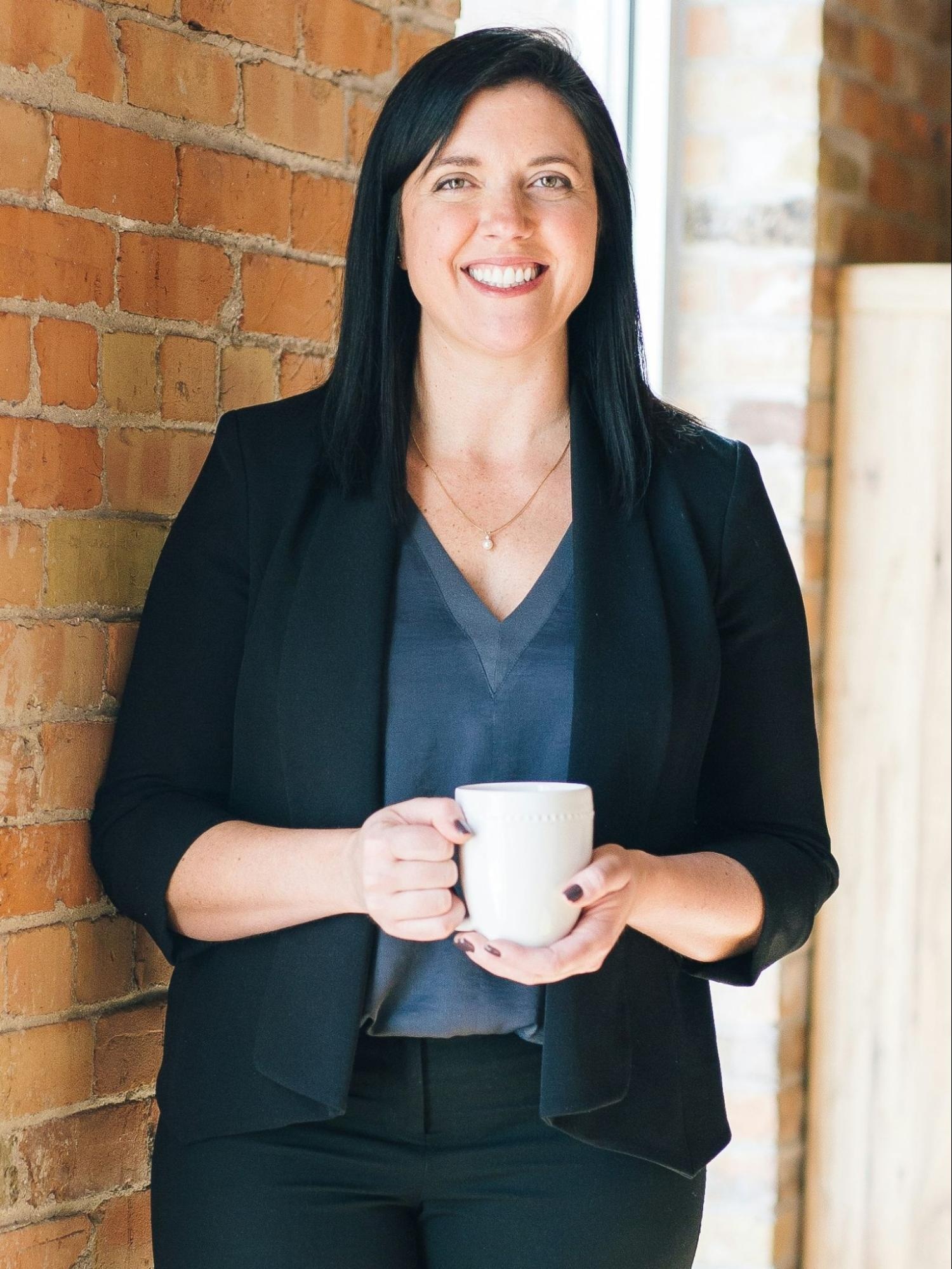 Mobile-friendly portrait crop of a smiling woman holding a white mug, standing by a brick wall with soft natural light in a professional indoor setting.