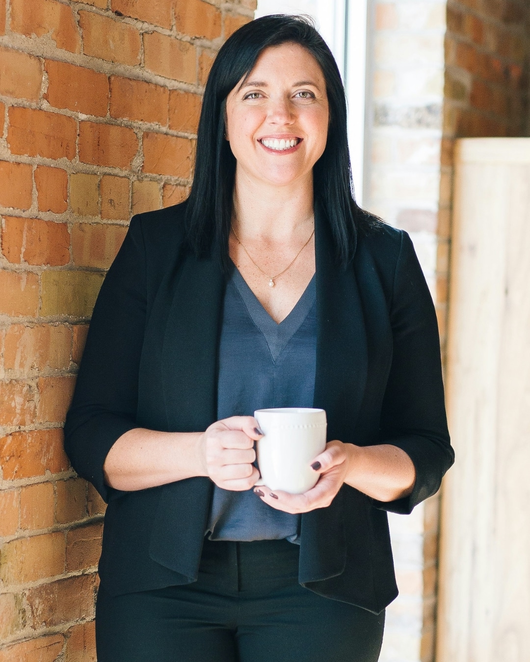 Portrait crop of a smiling woman holding a white mug, standing against an exposed brick wall in a bright, professional indoor space.