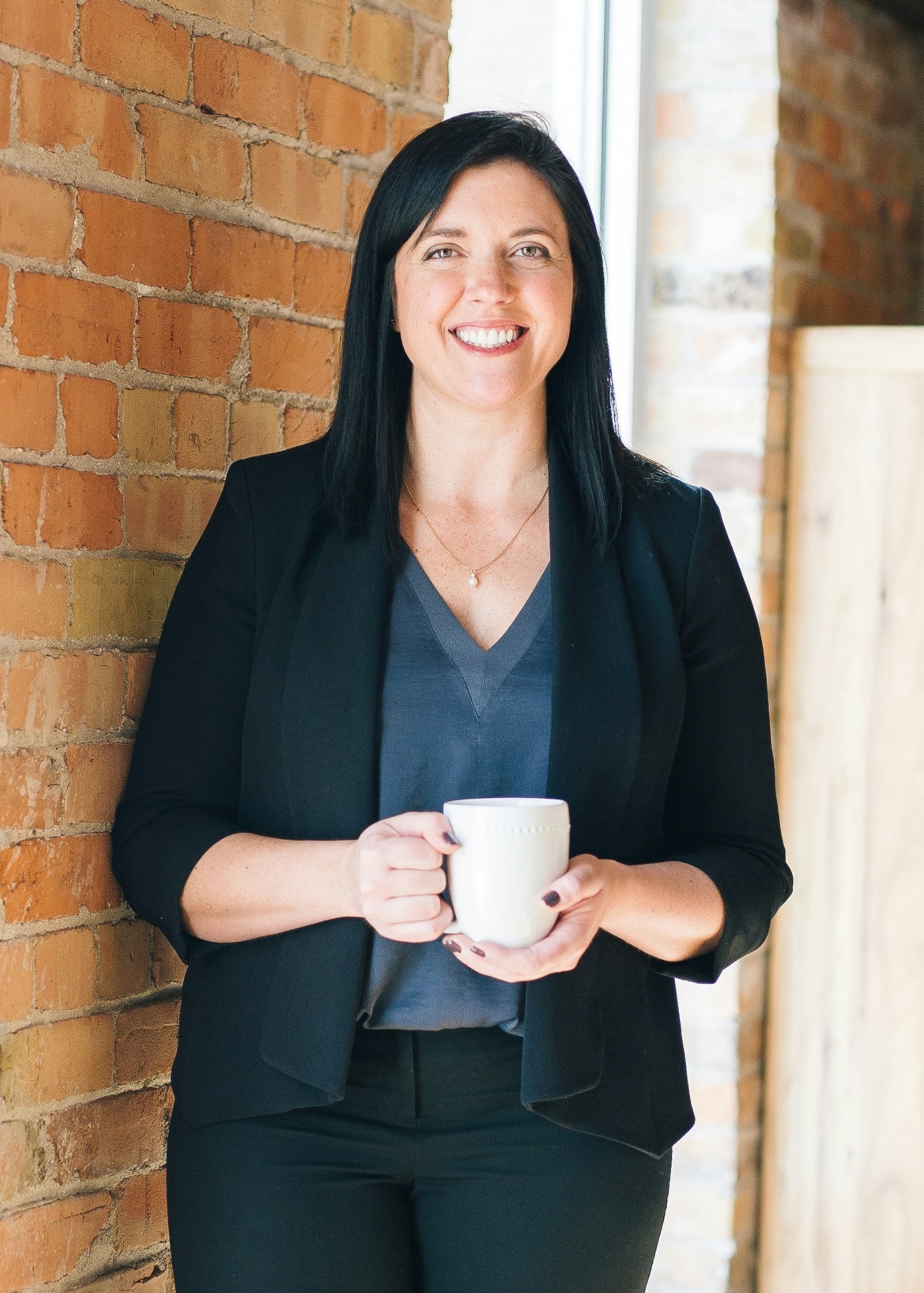 Professional photo print crop of a smiling woman holding a white mug, standing beside an exposed brick wall with natural window light in an office setting.
