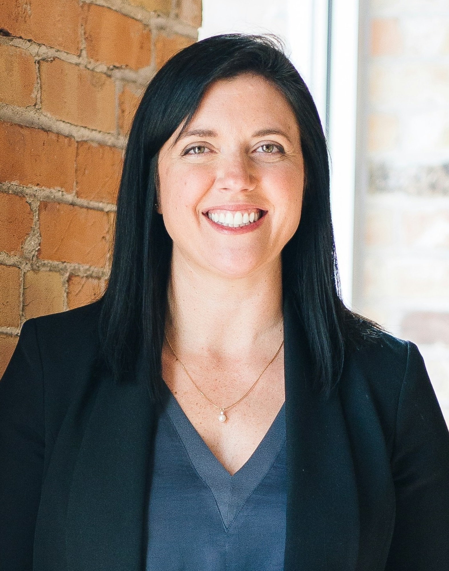 US visa and government ID crop of a smiling woman in business attire, positioned against a brick wall with soft window light.