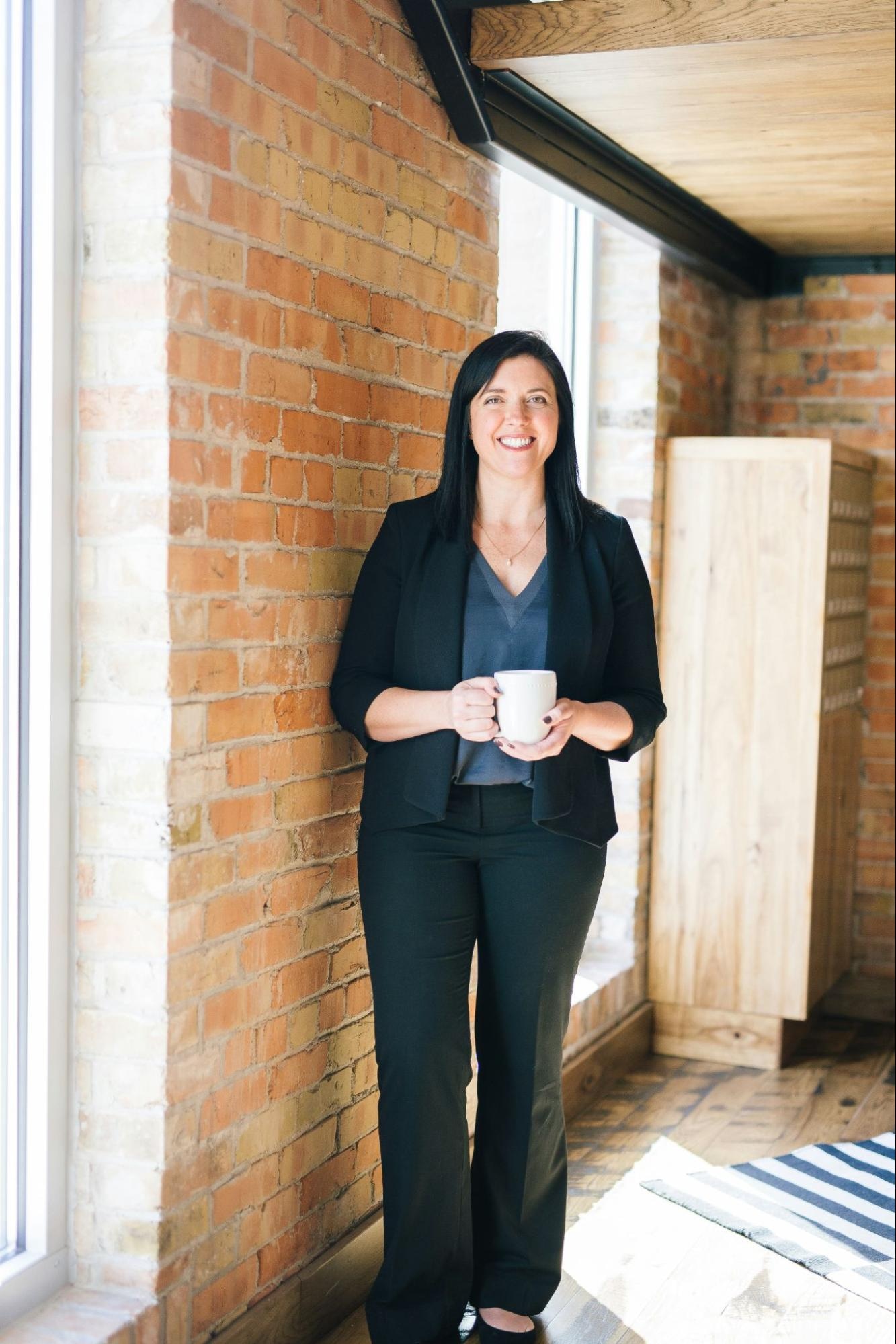 Businesswoman standing with a mug near a brick wall and sunlit window inside a modern office space.