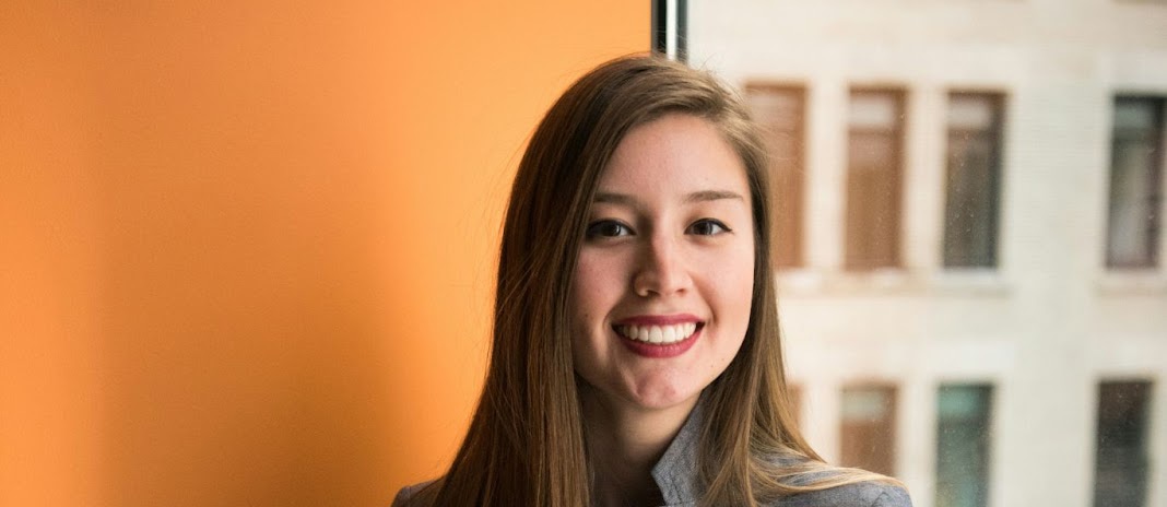 Panoramic crop of a smiling woman by a large window and vibrant orange wall, with soft daylight and an urban backdrop.
