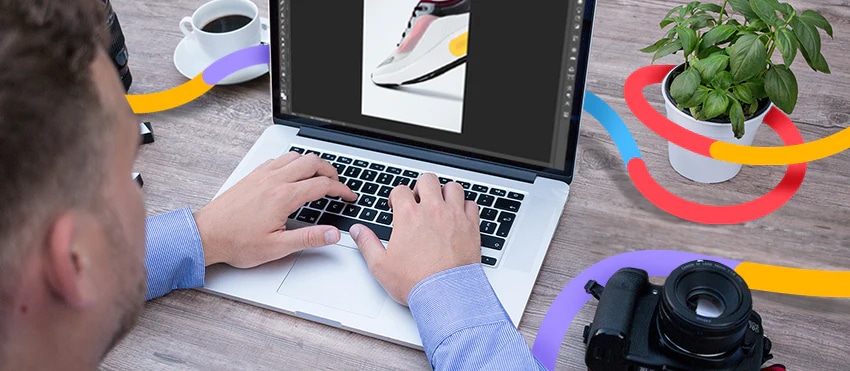Person editing a shoe photo on laptop with camera, coffee, and plant on workspace.