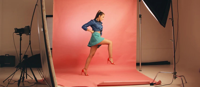 Female model in denim jacket and teal skirt posing in front of a pink backdrop in a professional photo studio.