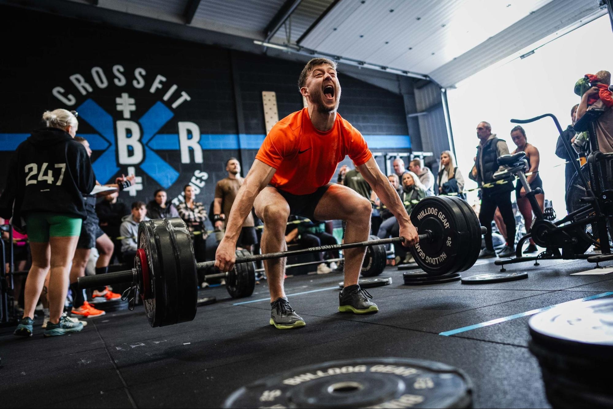 Man in orange shirt lifting heavy weights in a CrossFit gym, surrounded by spectators and gym equipment