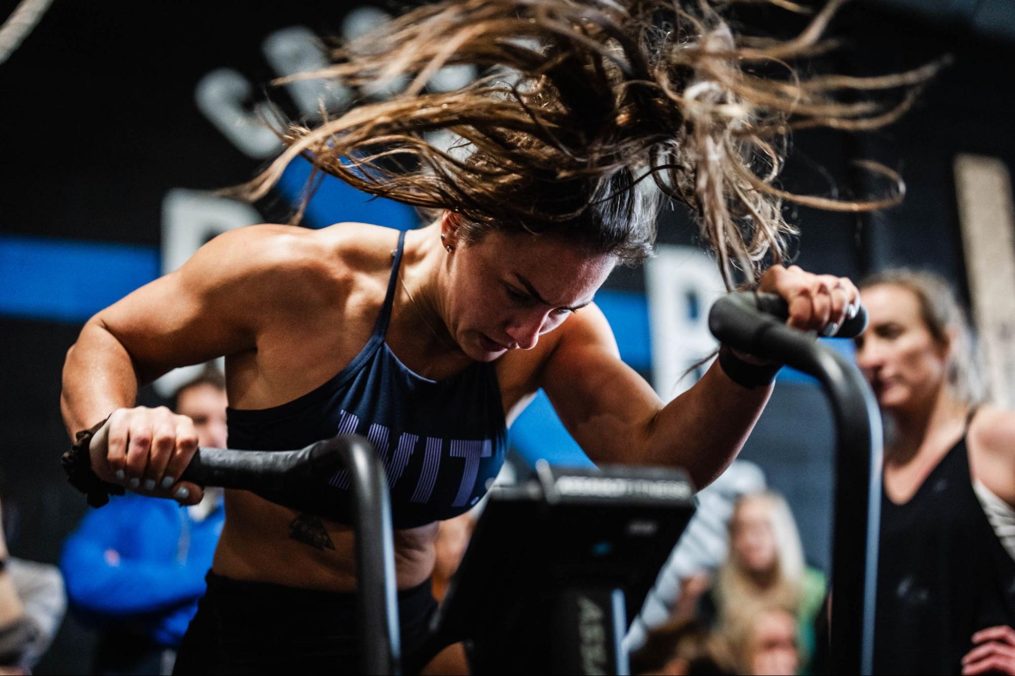 Woman with flowing hair intensely working out on an assault bike at a CrossFit gym, surrounded by other gym-goers