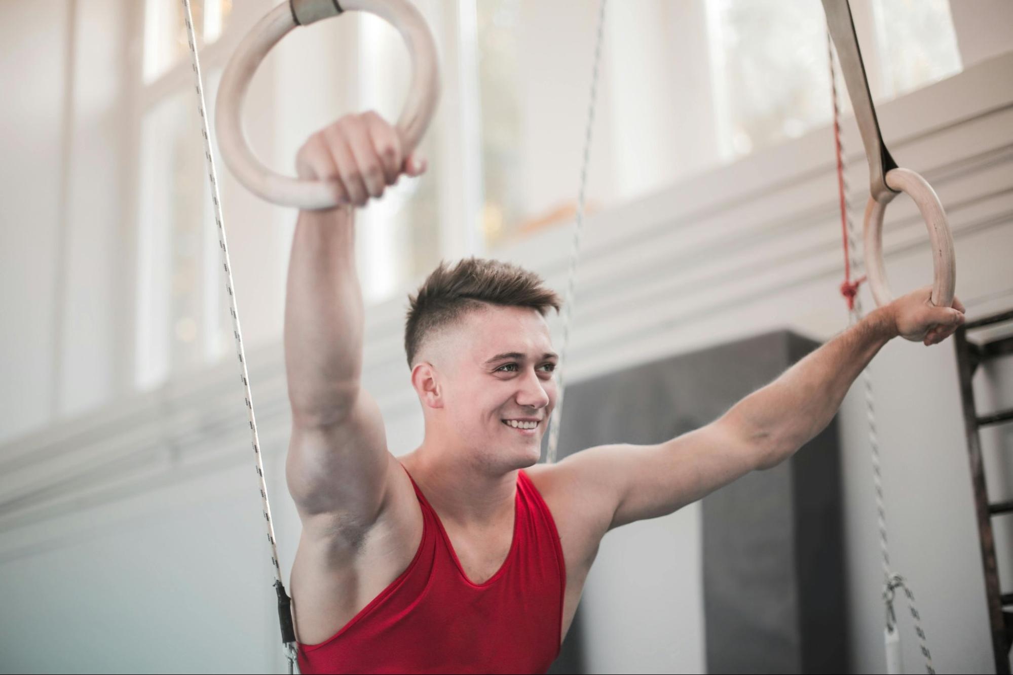 Smiling male gymnast in a red tank top holding gymnastics rings during training session in gym.