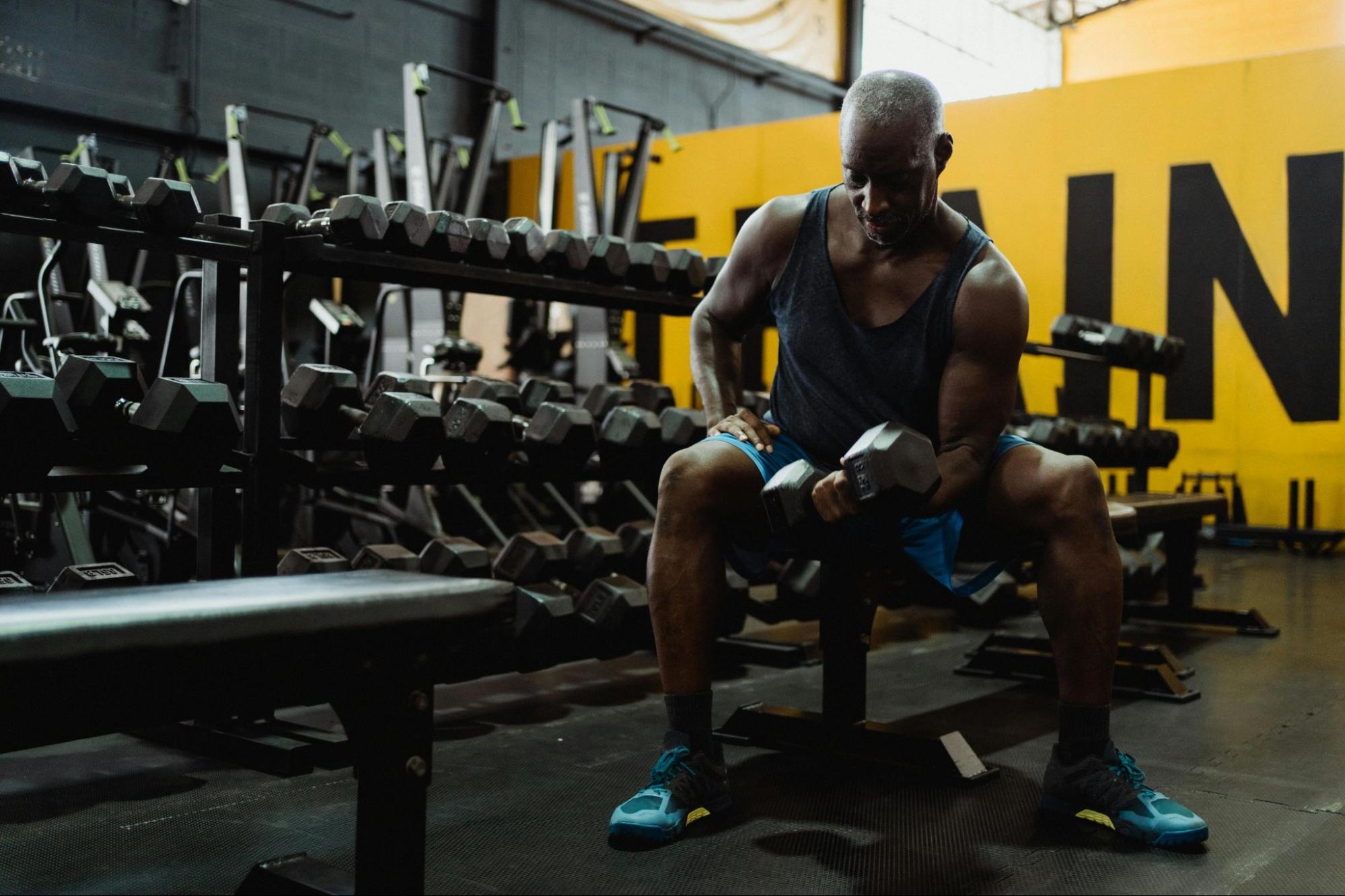 Older man lifting a dumbbell in a gym, seated in front of a rack of weights, focused on his workout.