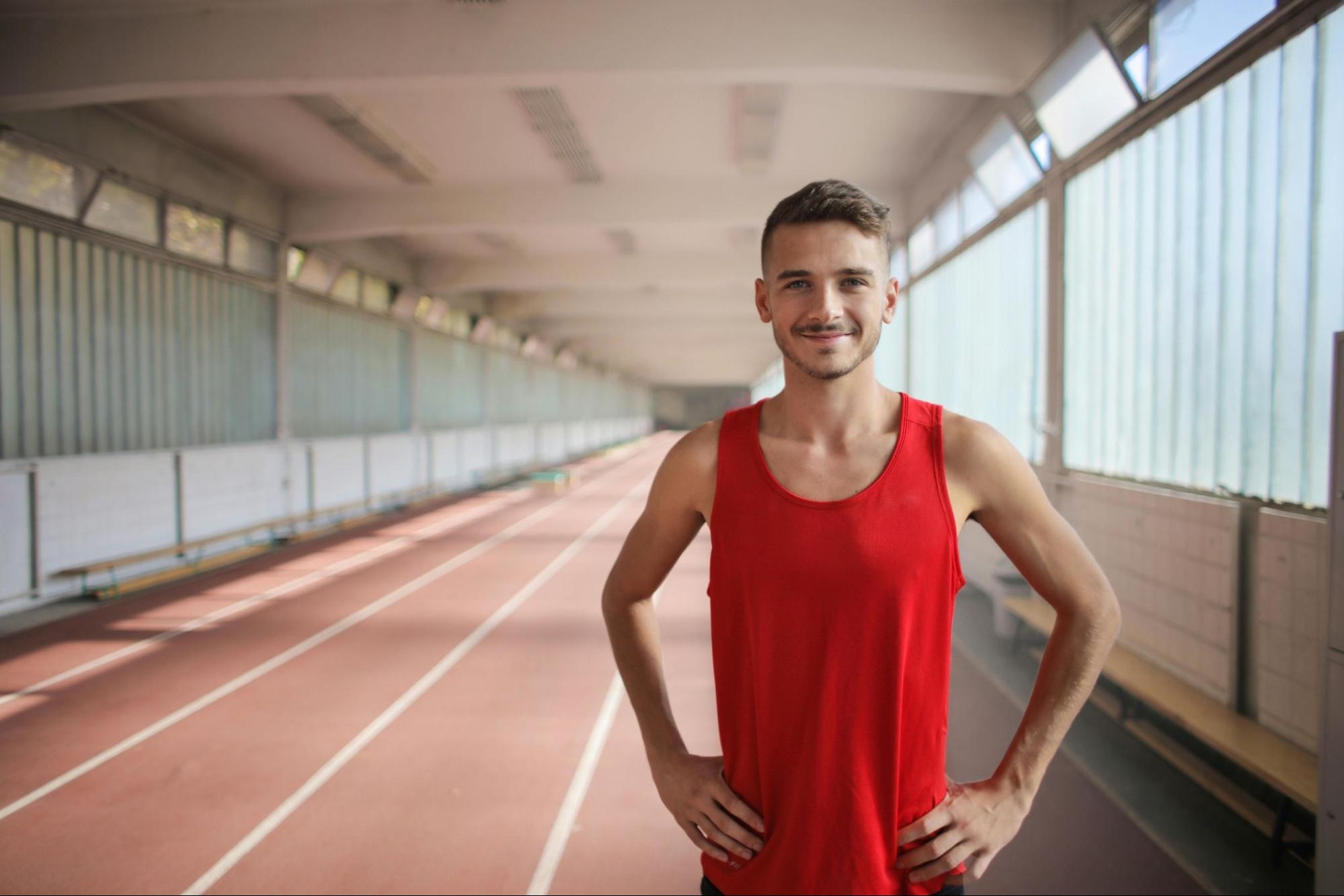 A close-up portrait of a male track athlete standing on an indoor track, wearing a red tank top.