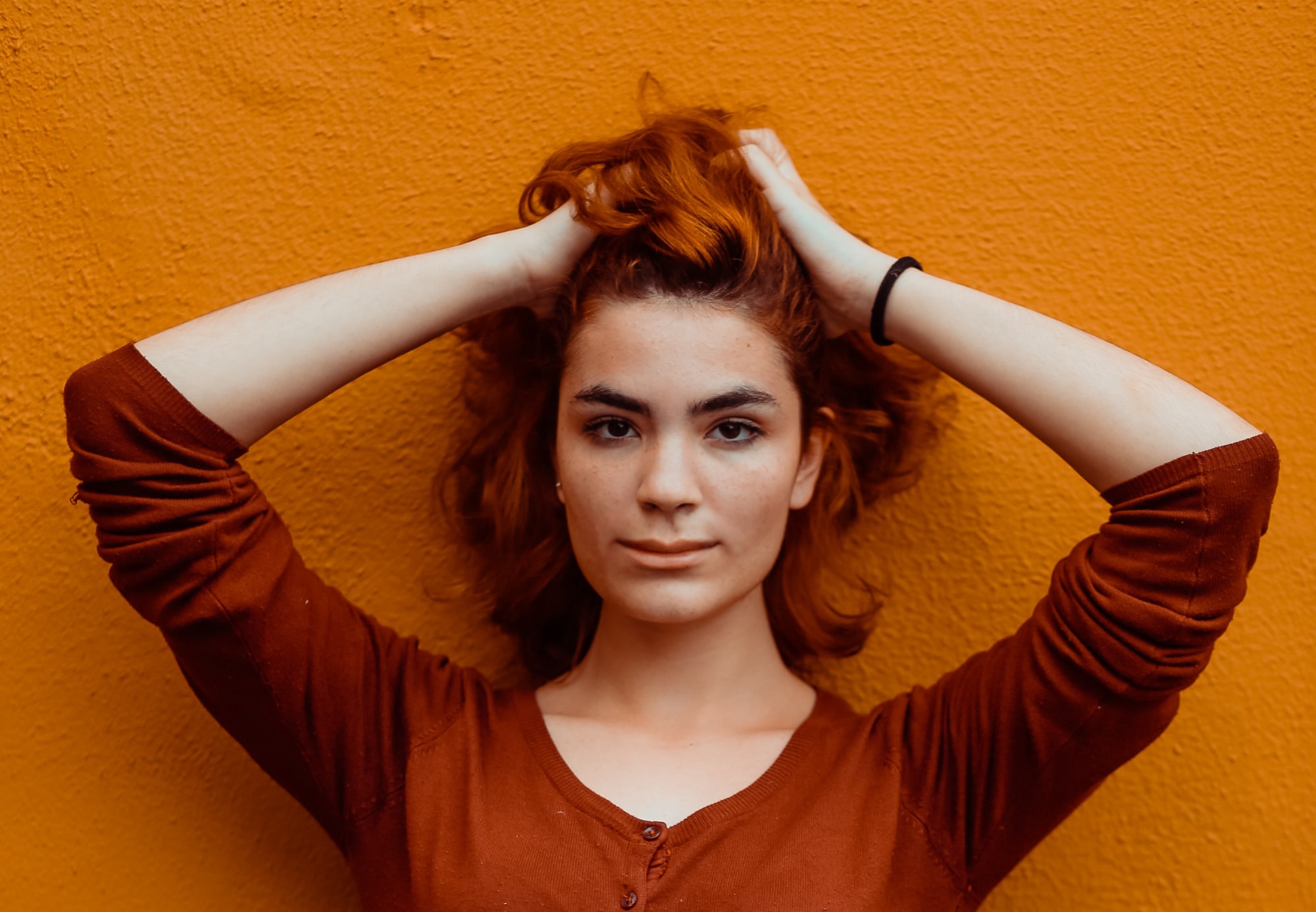 Portrait of a woman standing against an orange background with hands in her hair.