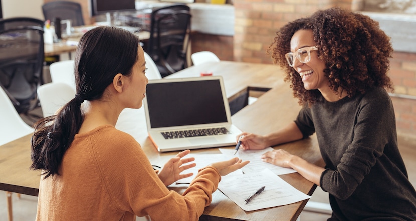 Two women engaged in a friendly business discussion at a desk with a laptop and documents.