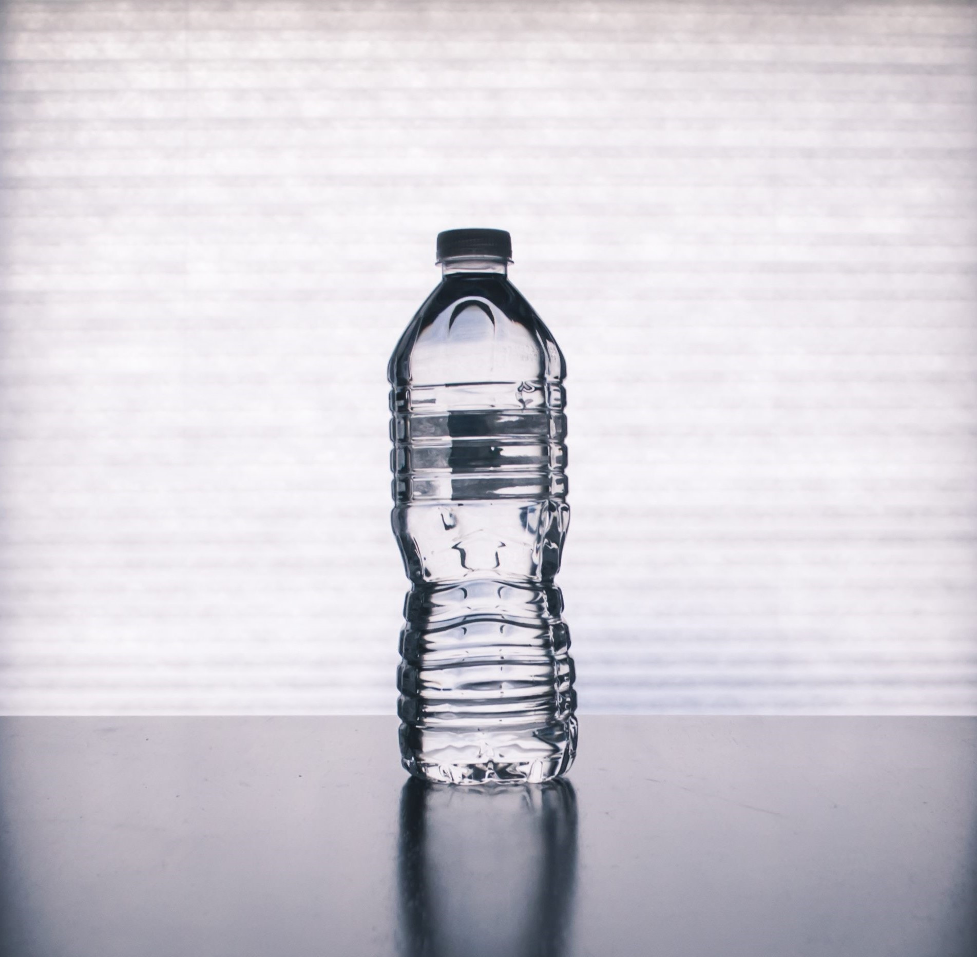 Clear plastic water bottle with cap, placed on a reflective surface against a bright, blurred background.
