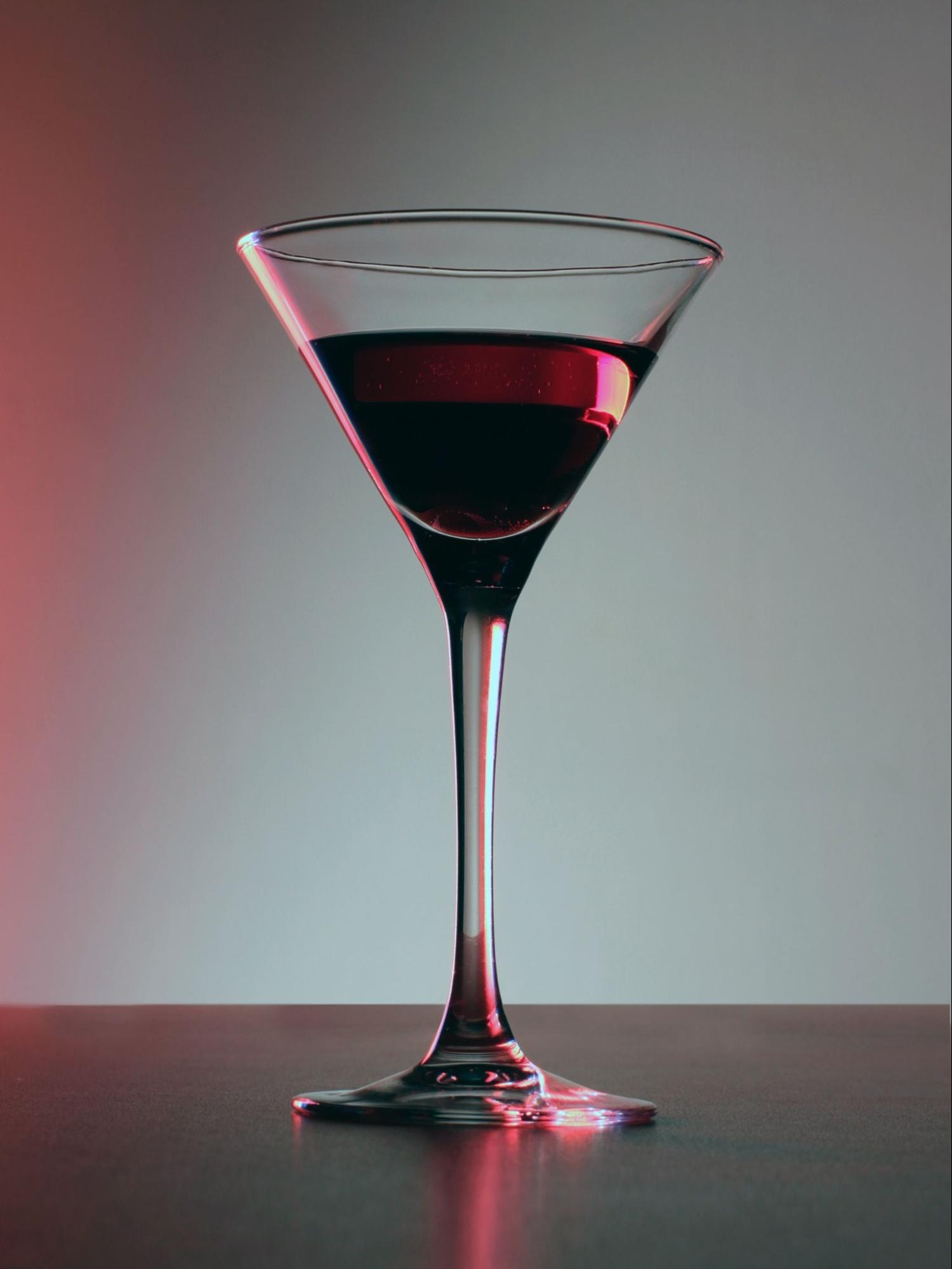 Martini glass filled with red wine on a dark surface, illuminated by gradient red and gray lighting.