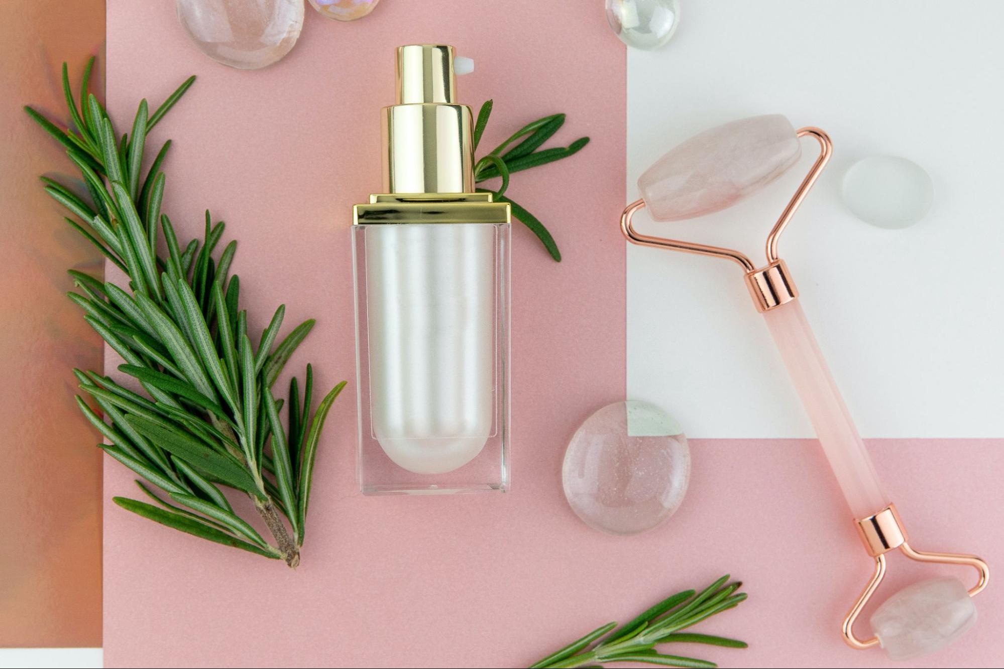 Skincare serum bottle with jade roller and rosemary on pink background, showcasing natural beauty products.