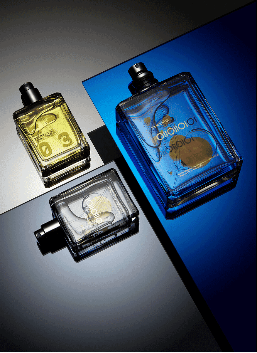 Three glass perfume bottles in yellow, blue, and clear displayed on reflective surfaces with dramatic lighting