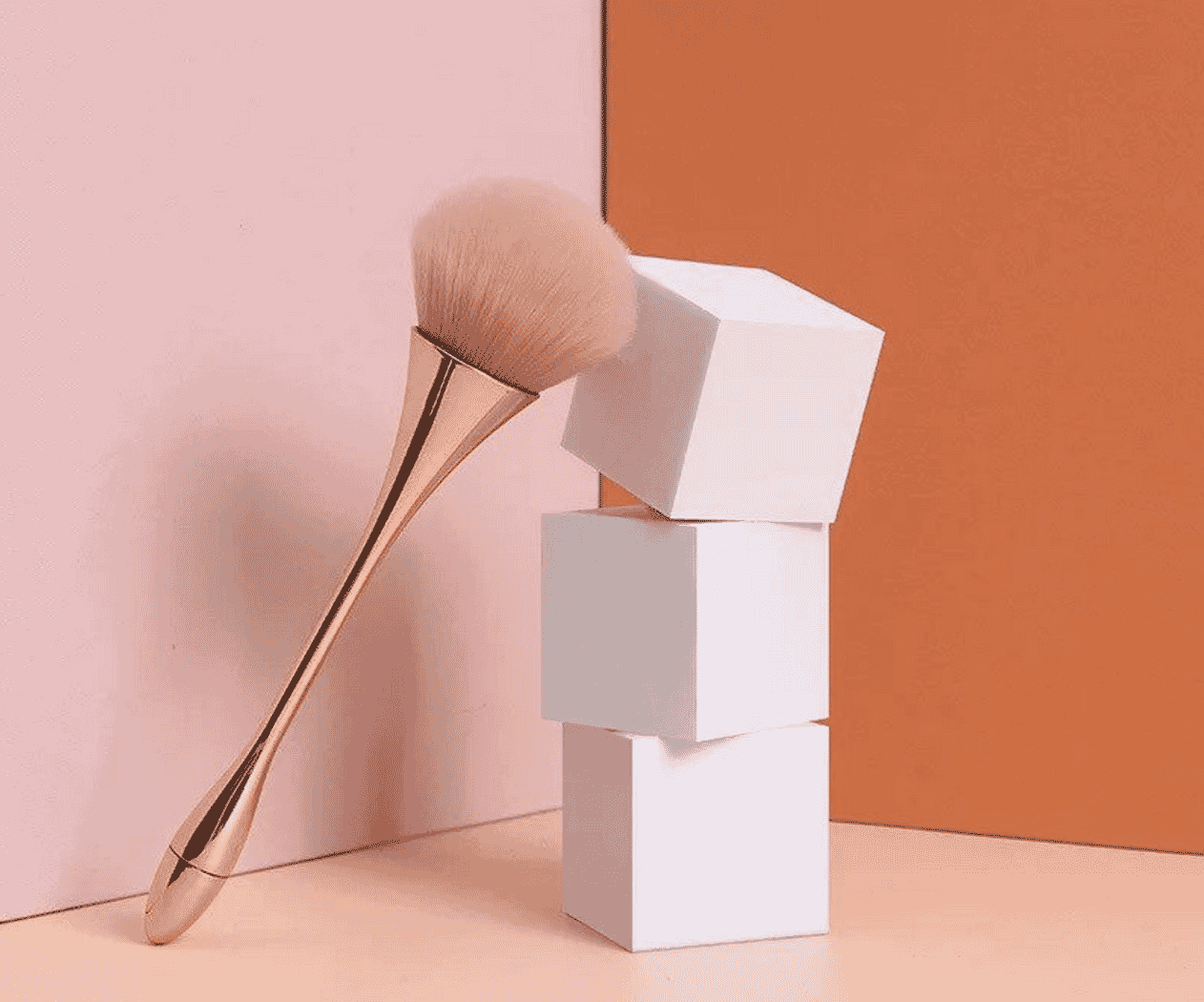 Fluffy makeup brush with gold handle leaning on stacked white cubes against pink and orange background
