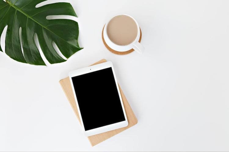 Flat lay of a tablet on a notebook, a coffee cup, and a large Monstera leaf on a white background.