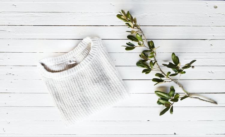 Flat lay of folded white sweater with an olive branch on white wooden background, styled minimalistically.