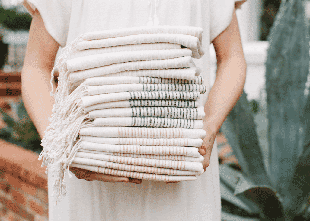 Person holding a stack of folded, handwoven textiles in neutral and striped patterns outdoors.