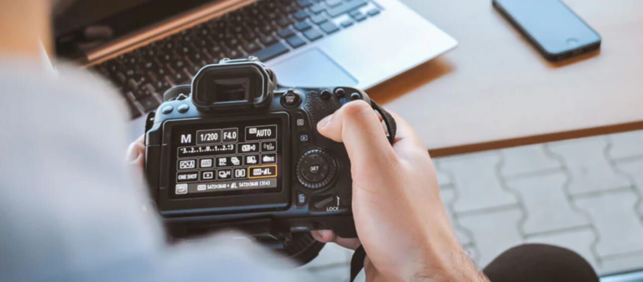 Photographer holding DSLR camera, adjusting settings with laptop and smartphone on nearby table.