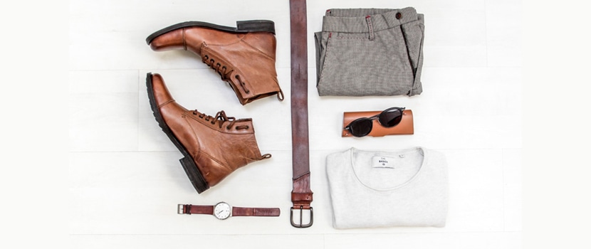 Flat lay of men’s travel essentials: boots, belt, watch, pants, shirt, and sunglasses