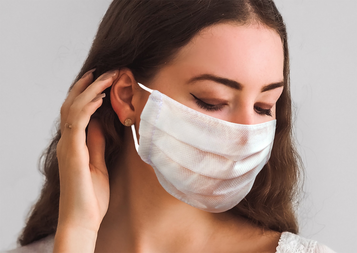 Close-up of a woman wearing a white face mask, adjusting the ear strap with her hand.