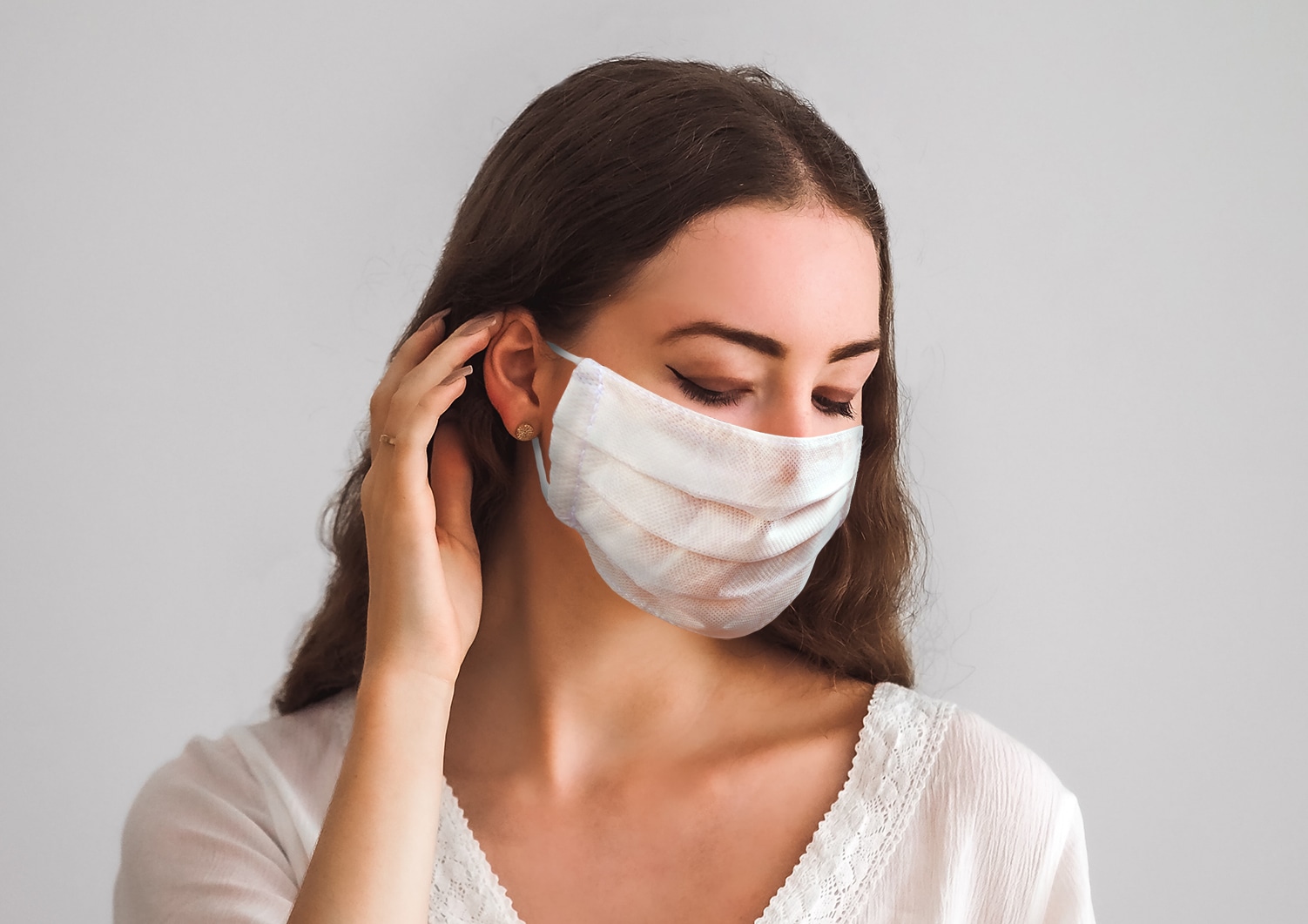 Woman with long hair wearing a white face mask, looking down with her hand near her ear.