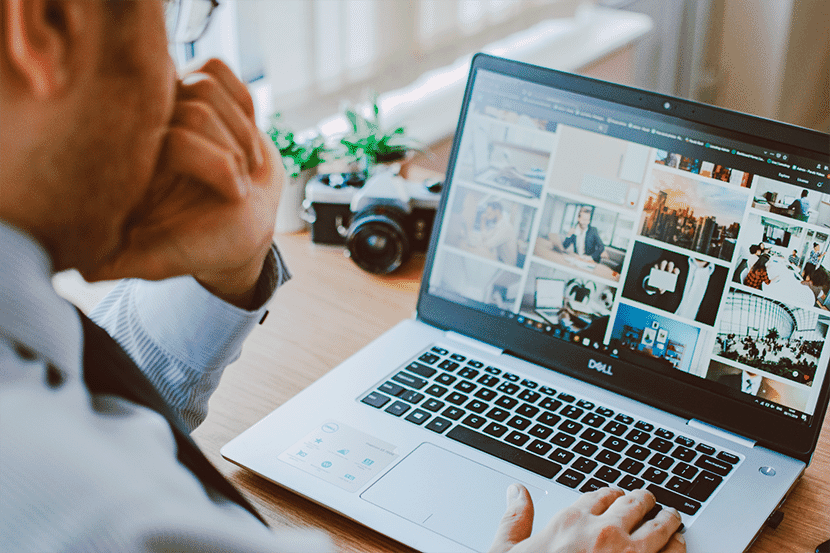 Man working on a laptop, reviewing photo thumbnails with a camera and plant on the desk.