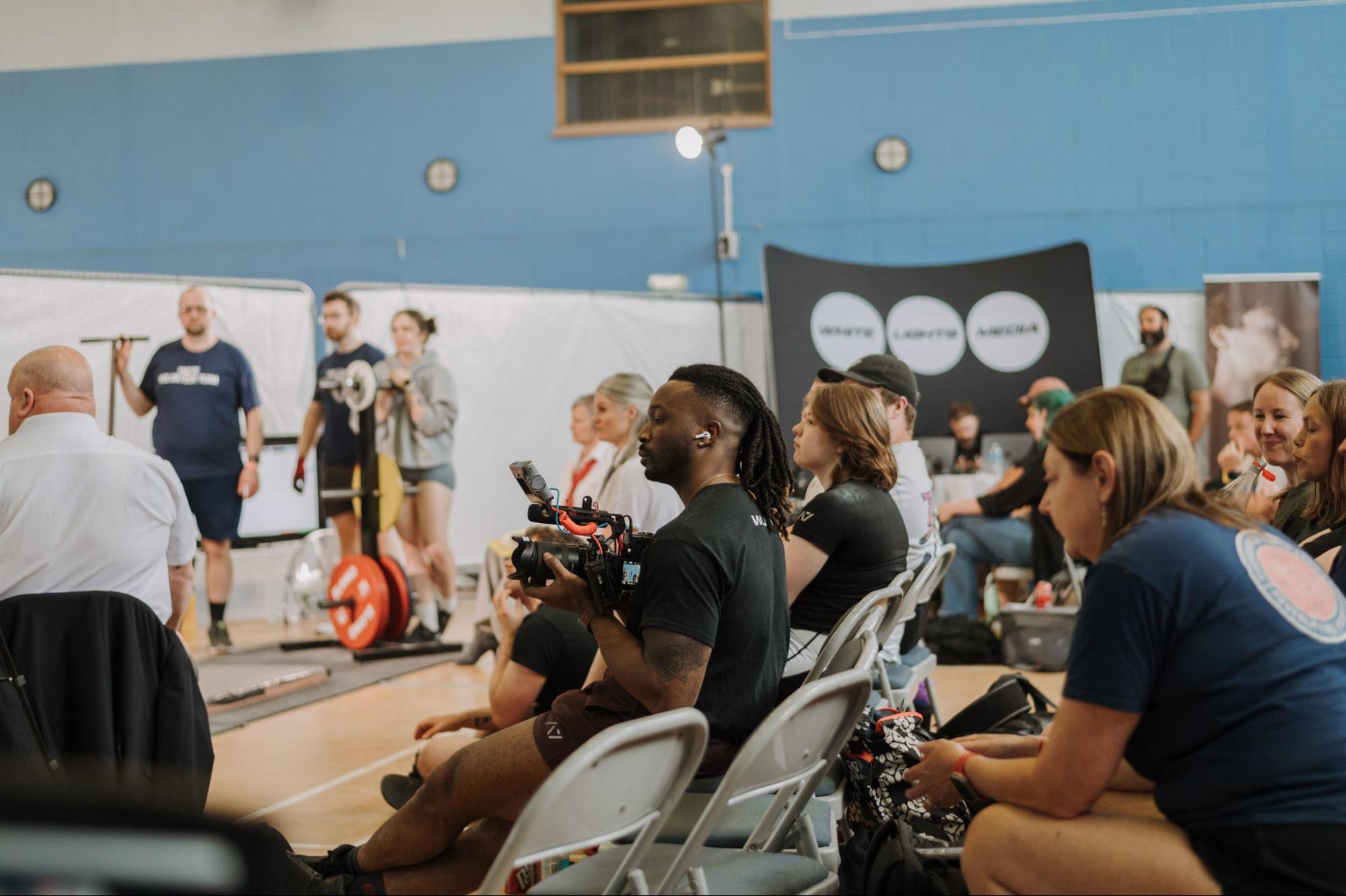 Photographer seated with camera in hand at a sports event, surrounded by audience members and athletes