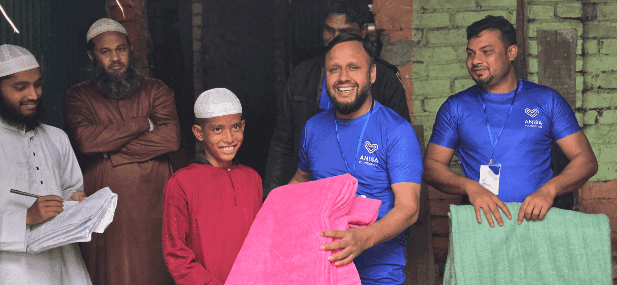 man smiling and giving a blanket to a smiling boy, with other adult men in the background